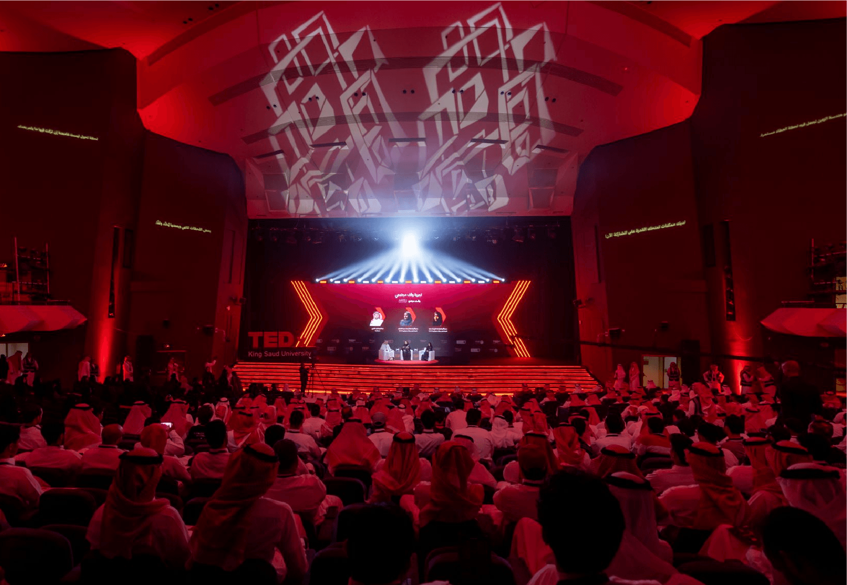 Full auditorium at TEDx King Saud University, red stage lighting, geometric ceiling projection, and a packed crowd